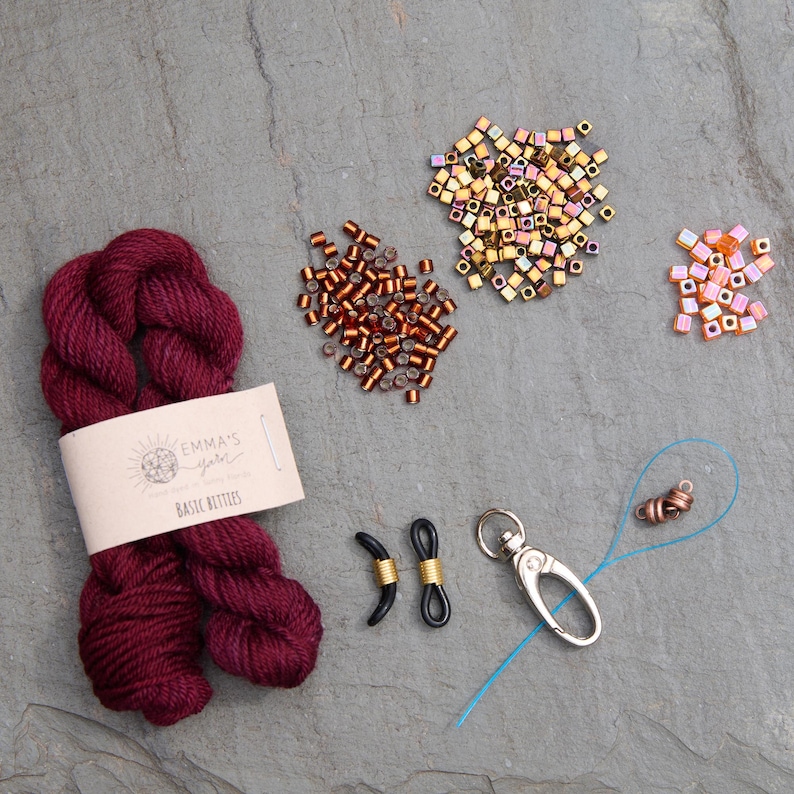 Pu&ograve; includere: Una collezione di materiali per l'artigianato, tra cui un gomitolo di lana bordeaux con l'etichetta "Emma's Yarns Basic Bettles", varie perline, un fermaglio argentato e fermi per occhiali neri con filo dorato.