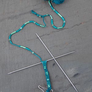 Pu&ograve; includere: Un gomitolo di filato verde acqua con un filo di perline e ferri da maglia. Il filo di perline &egrave; verde acqua e verde. I ferri da maglia sono argentati. Un fermaglio argentato &egrave; attaccato al pezzo lavorato a maglia.