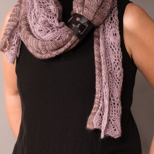 May include: A woman wearing a black sleeveless top with a delicate, light purple and brown knitted scarf. The scarf is secured with a black leather buckle.
