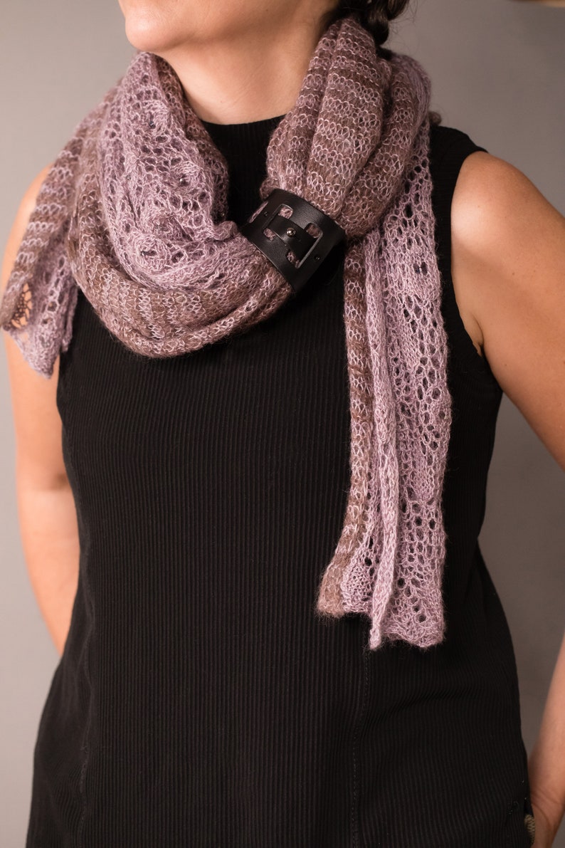 May include: A woman wearing a black sleeveless top with a delicate, light purple and brown knitted scarf. The scarf is secured with a black leather buckle.