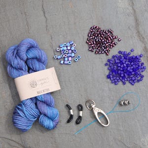 Pu&ograve; includere: Un gomitolo di filo blu con un'etichetta che recita "Emma's Yarn BASIC BITTIES" &egrave; accanto a piccoli mucchi di perline viola e blu. Ci sono anche fermi per occhiali neri e un fermaglio argentato.