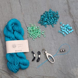 Pu&ograve; includere: Un assortimento di materiali per l'artigianato, tra cui un gomitolo di filato turchese con l'etichetta "Emma's Yarn Basic Bitties", varie perline turchesi e verdi, fermi per occhiali neri e un moschettone argentato.