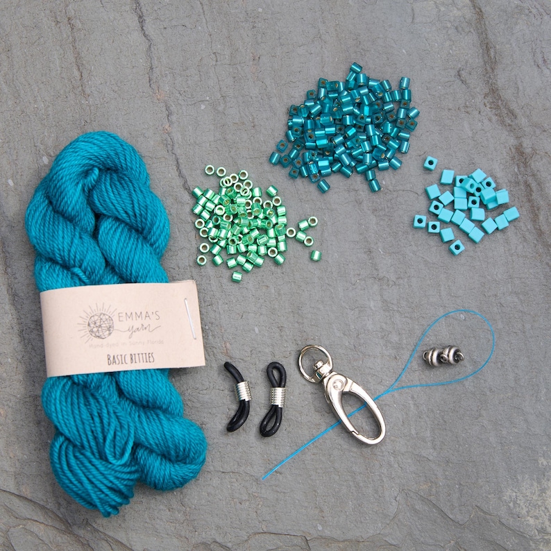 Pu&ograve; includere: Un assortimento di materiali per l'artigianato, tra cui un gomitolo di filato turchese con l'etichetta "Emma's Yarn Basic Bitties", varie perline turchesi e verdi, fermi per occhiali neri e un moschettone argentato.