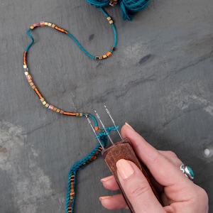 May include: A wooden knitting fork tool with two metal prongs being used to weave teal yarn. The yarn is threaded with small, metallic, square-shaped beads in shades of copper, bronze, and gold, creating a decorative, beaded cord design.