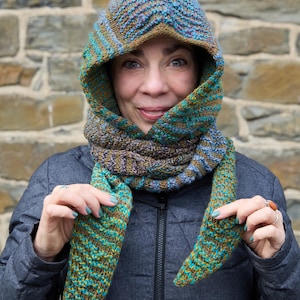 Puede incluir: Bufanda con capucha de punto en tonos turquesa, marrón y azul. La bufanda tiene capucha y extremos largos. La persona de la imagen lleva una chaqueta gris oscuro. El fondo es una pared de piedra.