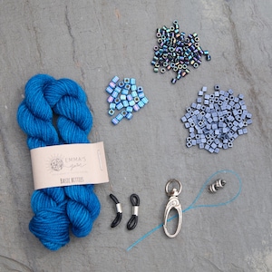Pu&ograve; includere: Una vista dall'alto di materiali per l'artigianato su una superficie grigia. Un gomitolo di filato blu brillante con un'etichetta che recita "Emma's Yarn Basic Betties" &egrave; accanto a piccoli mucchi di perline iridescenti, fermi per occhiali neri e un fermaglio argentato.