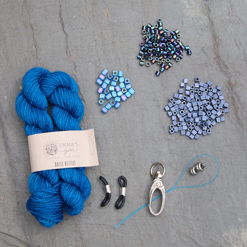 Pu&ograve; includere: Una vista dall'alto di materiali per l'artigianato su una superficie grigia. Un gomitolo di filato blu brillante con un'etichetta che recita "Emma's Yarn Basic Betties" &egrave; accanto a piccoli mucchi di perline iridescenti, fermi per occhiali neri e un fermaglio argentato.
