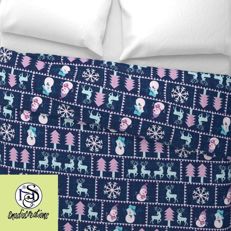 May include: A navy blue fabric with a repeating pattern of pink trees, white snowflakes, blue reindeer, and white snowmen with pink hats. The fabric is folded in half.