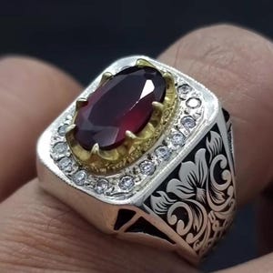 May include: A silver ring with a large, oval-shaped, dark red gemstone set in a gold bezel. The ring is decorated with smaller, clear gemstones around the bezel and intricate floral designs on the band.