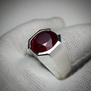 Könnte beinhalten: Ein Silberring mit einem großen, oval geschliffenen Rubin-Edelstein. Der Rubin hat eine tiefrote Farbe und ist in einer geometrischen Lünette gefasst. Der Ring hat ein breites Band und eine polierte Oberfläche.