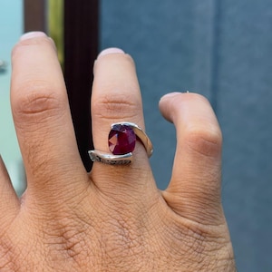 May include: A silver bypass ring featuring a faceted oval-cut deep red gemstone set between two open ends. The band displays a detailed black engraved scrollwork pattern along the outer surface, creating a distinct contrast against the polished silver metal.