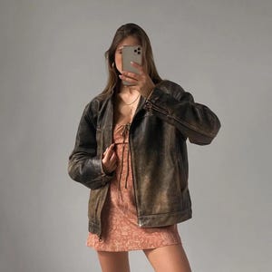 May include: A distressed brown leather jacket with a zip-up front. The jacket is worn over a peach-colored dress with a floral pattern. The outfit is set against a plain gray background.