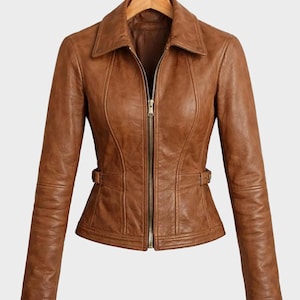 Könnte beinhalten: Eine hellbraune Lederjacke mit spitzem Kragen, durchgehendem Reißverschluss in der Mitte und langen Ärmeln. Die Jacke verfügt über seitliche Taillenverstelllaschen mit Schnallen und vertikale Nahtdetails für eine taillierte Passform.