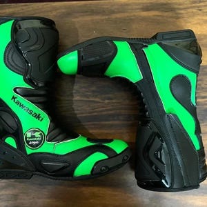 Könnte beinhalten: Paar schwarze und grüne Kawasaki Motorradstiefel. Die Stiefel haben ein hohes Design mit grünen Akzenten und dem Kawasaki-Logo. Die Stiefel bestehen aus einem strapazierfähigen Material und sind auf Schutz und Leistung ausgelegt.