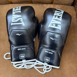 May include: A pair of black Everlast boxing gloves with white laces. The gloves feature the white Everlast logo, the text EST.1910, and a small square patch with the text HECHO EN MEXICO and 20 OZ printed on the wrist area of each glove.