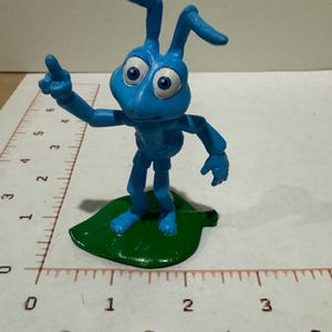 Può includere: Una statuetta di formica blu su una base a forma di foglia verde, posizionata accanto a un righello che mostra un'altezza di circa 9 cm. La formica ha grandi occhi bianchi con pupille blu, due antenne e il braccio destro sollevato.