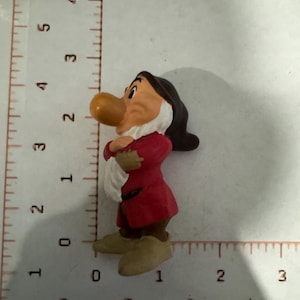 Pode incluir: Uma pequena estatueta do Zangado da Branca de Neve e os Sete Anões, vestindo uma túnica vermelha, calças castanhas e sapatos bege, ao lado de uma régua que mostra uma altura de cerca de 6,4 cm e uma largura de 3,8 cm.