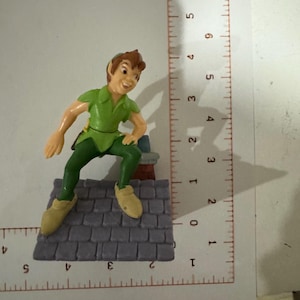 May include: A figurine of Peter Pan, dressed in a green tunic and pants, with tan shoes, perched on a gray rooftop. The figure is approximately 3 inches tall.