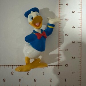May include: A small, colorful Donald Duck figurine. The figure is wearing a blue sailor shirt and hat, a red bow tie, and has a yellow beak and feet. The figurine is approximately 2 inches tall.