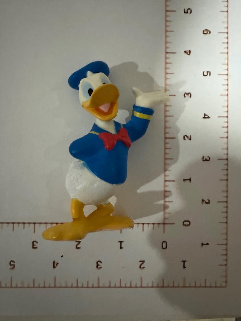 May include: A small, colorful Donald Duck figurine. The figure is wearing a blue sailor shirt and hat, a red bow tie, and has a yellow beak and feet. The figurine is approximately 2 inches tall.