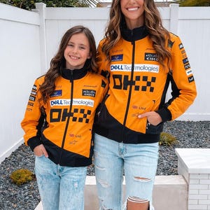 May include: Two people wearing matching orange and black racing jackets with sponsor logos and the text "DELL Technologies" and "DHX". They are also wearing light blue ripped jeans. The jackets have black accents and a black collar.