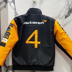 May include: Black McLaren Formula 1 Team jacket with orange sleeves and the number 4 in orange on the back. The jacket has a stand-up collar and various sponsor logos on the sleeves.