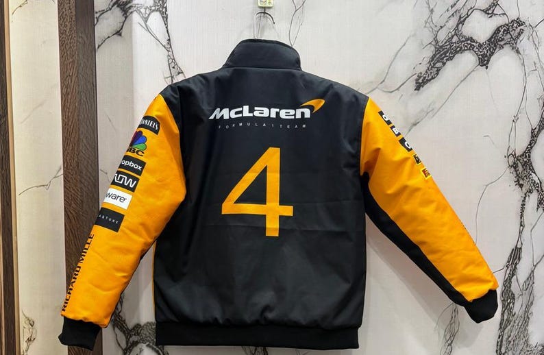May include: Black McLaren Formula 1 Team jacket with orange sleeves and the number 4 in orange on the back. The jacket has a stand-up collar and various sponsor logos on the sleeves.