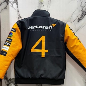 May include: Black McLaren Formula 1 Team jacket with orange sleeves and the number 4 in orange on the back. The jacket has a stand-up collar and various sponsor logos on the sleeves.