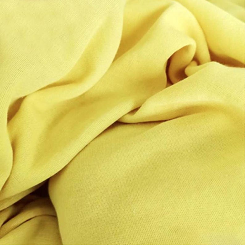 May include: Close-up of a soft, yellow fabric with visible texture and folds. The material appears to be lightweight and flowing, with varying shades of yellow due to the folds and shadows. The fabric is likely used for clothing or home decor.