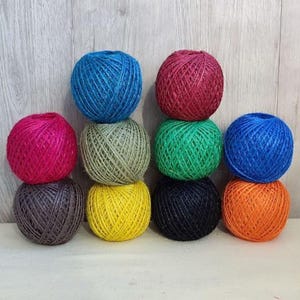 Op de afbeelding: Een verzameling van tien bollen touw in verschillende kleuren, waaronder blauw, rood, roze, groen, geel, zwart en oranje. De touwballen zijn gestapeld tegen een lichte houtnerf achtergrond. Het touw lijkt gemaakt van natuurlijke vezels.