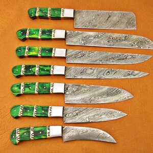 May include: A set of seven kitchen knives with patterned steel blades and green handles. The handles have a decorative design with gold-colored accents. The knives are arranged on a brown surface.