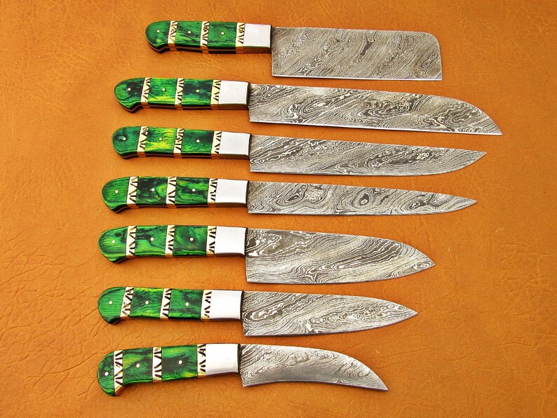May include: A set of seven kitchen knives with patterned steel blades and green handles. The handles have a decorative design with gold-colored accents. The knives are arranged on a brown surface.