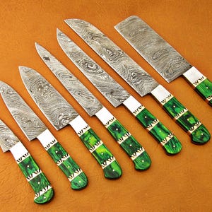 May include: An assortment of seven Damascus steel knives with green and gold handles. The blades have a unique patterned design. The handles are a vibrant green with gold accents. The knives are arranged on a brown surface.