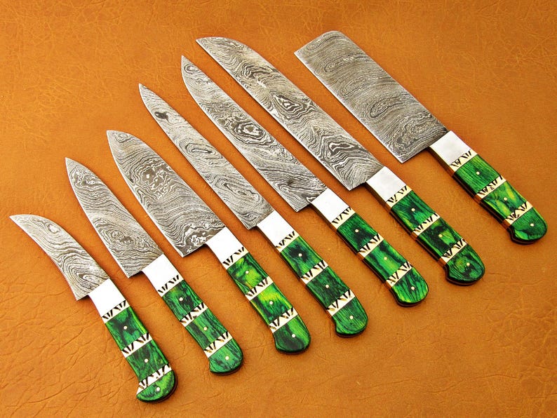 May include: An assortment of seven Damascus steel knives with green and gold handles. The blades have a unique patterned design. The handles are a vibrant green with gold accents. The knives are arranged on a brown surface.