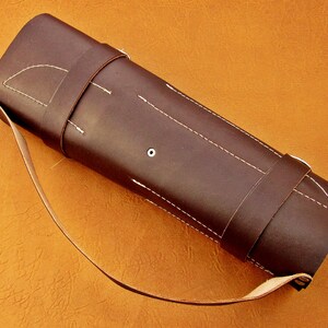 May include: A dark brown leather quiver with white stitching and a shoulder strap. The cylindrical case has a flap and a strap to secure the contents. The background is a solid orange color.