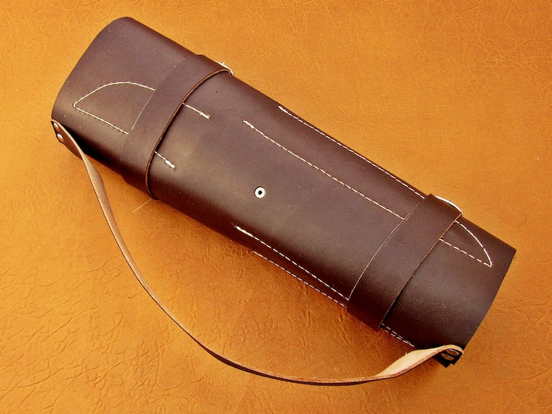 May include: A dark brown leather quiver with white stitching and a shoulder strap. The cylindrical case has a flap and a strap to secure the contents. The background is a solid orange color.