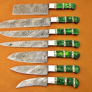 May include: Set of seven kitchen knives with patterned steel blades and green handles. The handles have a decorative design with gold-colored accents. The knives are arranged on a brown surface.