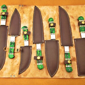 May include: A set of six knives with dark blades and green and white handles, each in a brown leather sheath. The knives are arranged on a tan leather roll with a buckle closure. The handles have a unique green and white pattern.