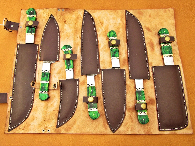 May include: A set of six knives with dark blades and green and white handles, each in a brown leather sheath. The knives are arranged on a tan leather roll with a buckle closure. The handles have a unique green and white pattern.
