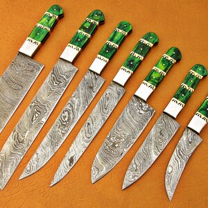 May include: An assortment of seven Damascus steel knives with green and gold handles. The blades have a unique swirling pattern. The handles are a vibrant green with gold accents. The knives are arranged diagonally on a brown surface.