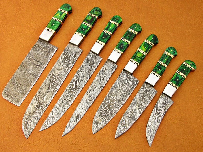 May include: An assortment of seven Damascus steel knives with green and gold handles. The blades have a unique swirling pattern. The handles are a vibrant green with gold accents. The knives are arranged diagonally on a brown surface.