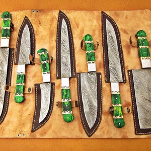 May include: A set of seven knives with patterned blades and green handles, displayed in a leather roll. The blades have a unique pattern, and the handles are accented with white and gold. The knives are secured with leather straps.