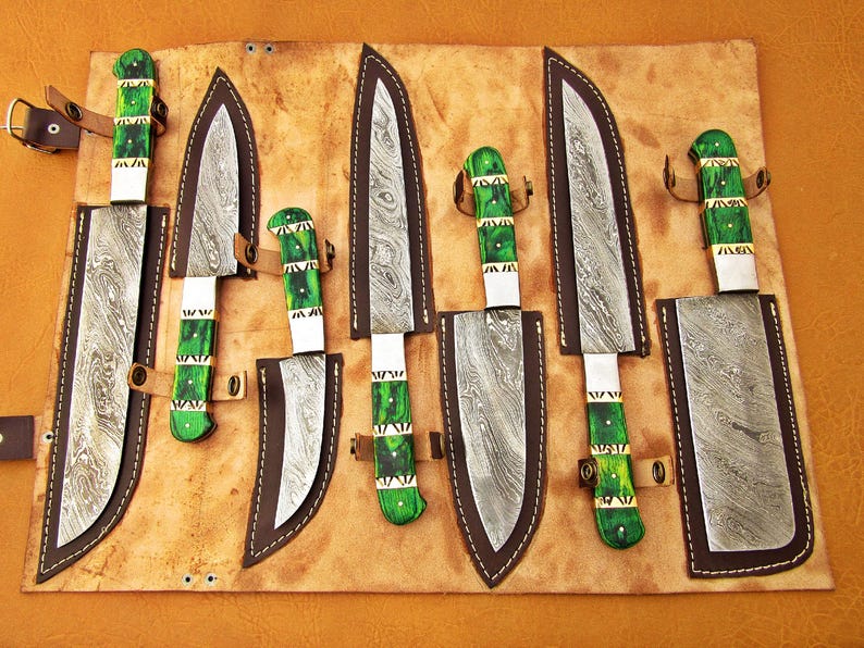 May include: A set of seven knives with patterned blades and green handles, displayed in a leather roll. The blades have a unique pattern, and the handles are accented with white and gold. The knives are secured with leather straps.