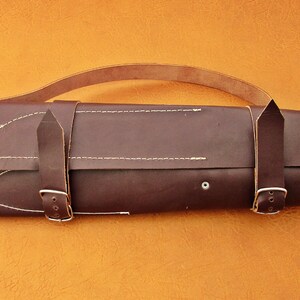 May include: A dark brown leather knife roll with a handle and two buckle straps. The roll is stitched with white thread and has a small silver rivet. The background is a solid orange color.