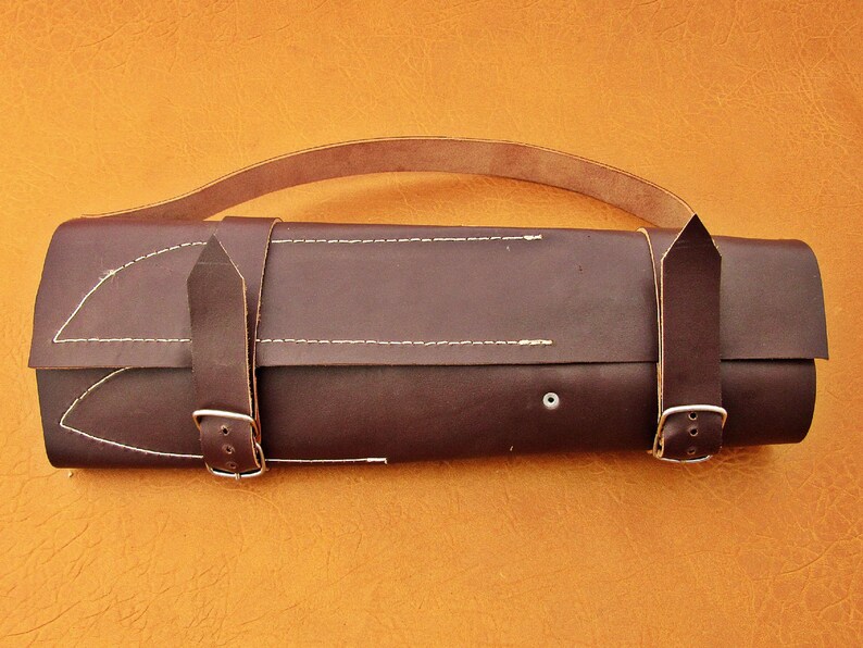 May include: A dark brown leather knife roll with a handle and two buckle straps. The roll is stitched with white thread and has a small silver rivet. The background is a solid orange color.