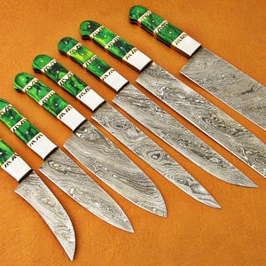 May include: A set of seven Damascus steel knives with green and white handles. The blades have a unique swirling pattern. The handles are a vibrant green with white accents. The knives are arranged diagonally on a brown surface.