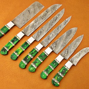 May include: Set of seven Damascus steel kitchen knives with green and gold handles. The blades have a unique patterned design. The handles are a vibrant green with gold accents. The knives are arranged on a brown surface.