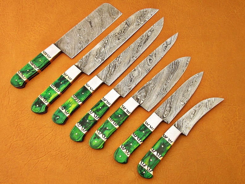 May include: Set of seven Damascus steel kitchen knives with green and gold handles. The blades have a unique patterned design. The handles are a vibrant green with gold accents. The knives are arranged on a brown surface.