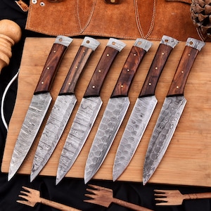 Puede incluir: Conjunto de seis cuchillos de cocina con mangos de madera y hojas de acero Damasco. Los cuchillos están dispuestos en una tabla de cortar de madera.