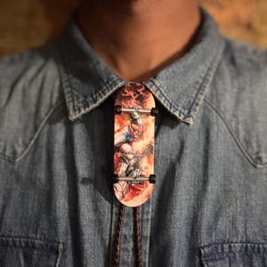 May include: A novelty bolo tie with a miniature skateboard design. The skateboard has a colourful graphic of a superhero. The tie is attached to a brown braided cord and is worn on a denim shirt.
